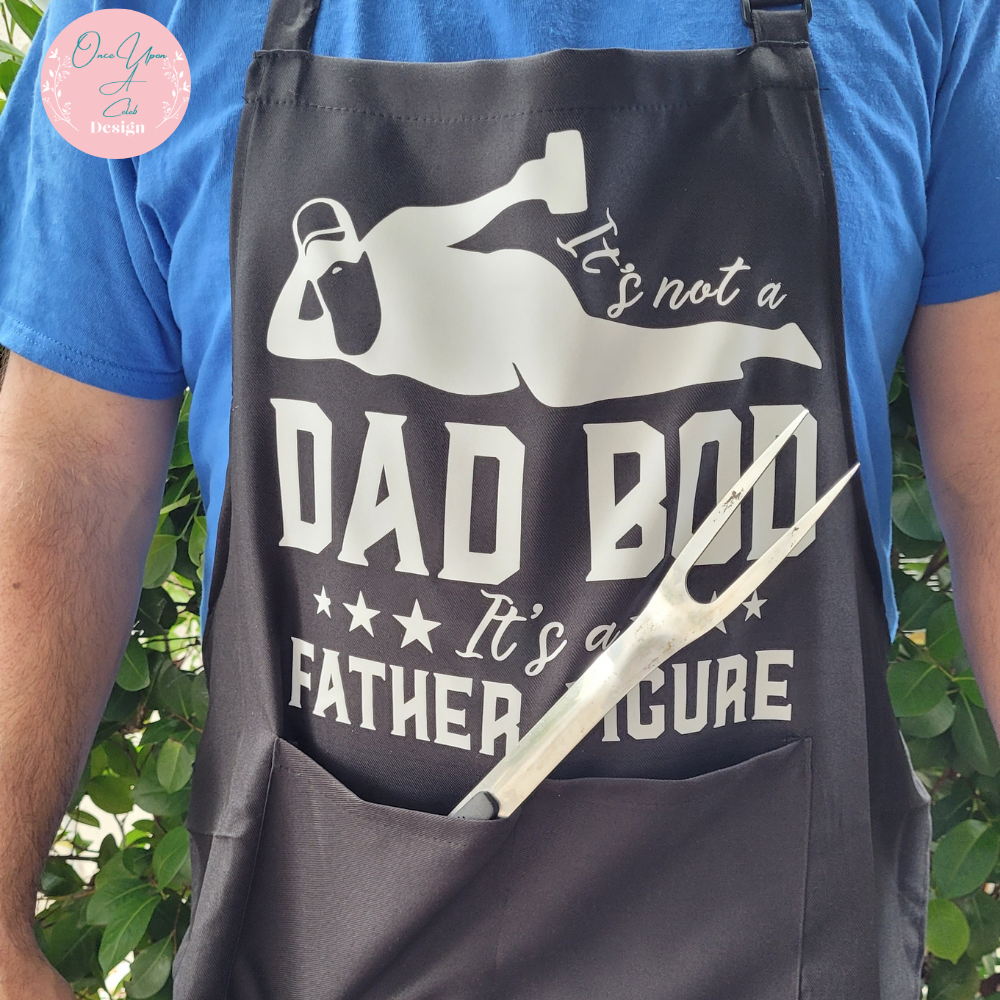 Black Apron With White Design of It's not a Dad Bod Its a Father Figure and showing A BBQ utensil in the front Pocket BY Once Upon A Celeb Design