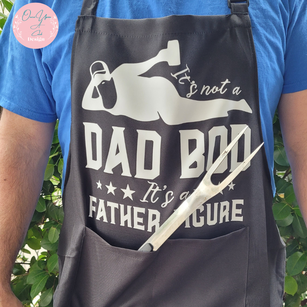 Black Apron With White Design of It's not a Dad Bod Its a Father Figure  and showing A BBQ utensil in the front Pocket BY Once Upon A Celeb Design